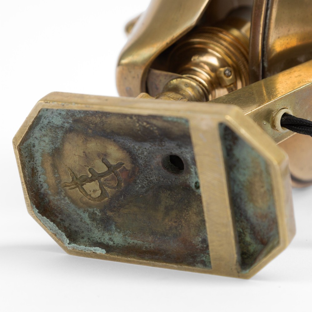 A polished brass table lamp featuring a stylised human-machine mask mounted in front of a circular dish reflector base. View IX