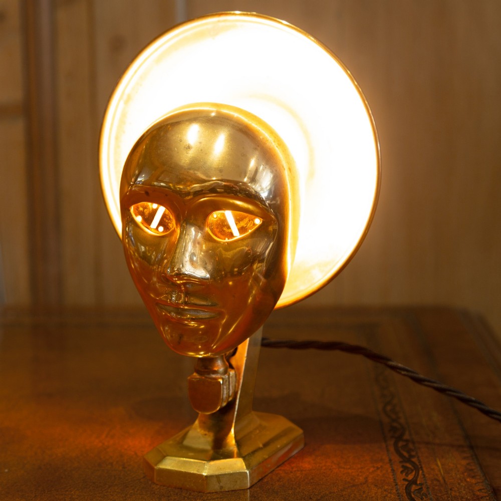 A polished brass table lamp featuring a stylised human-machine mask mounted in front of a circular dish reflector base. View X