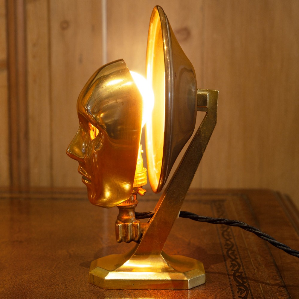 A polished brass table lamp featuring a stylised human-machine mask mounted in front of a circular dish reflector base. View XI