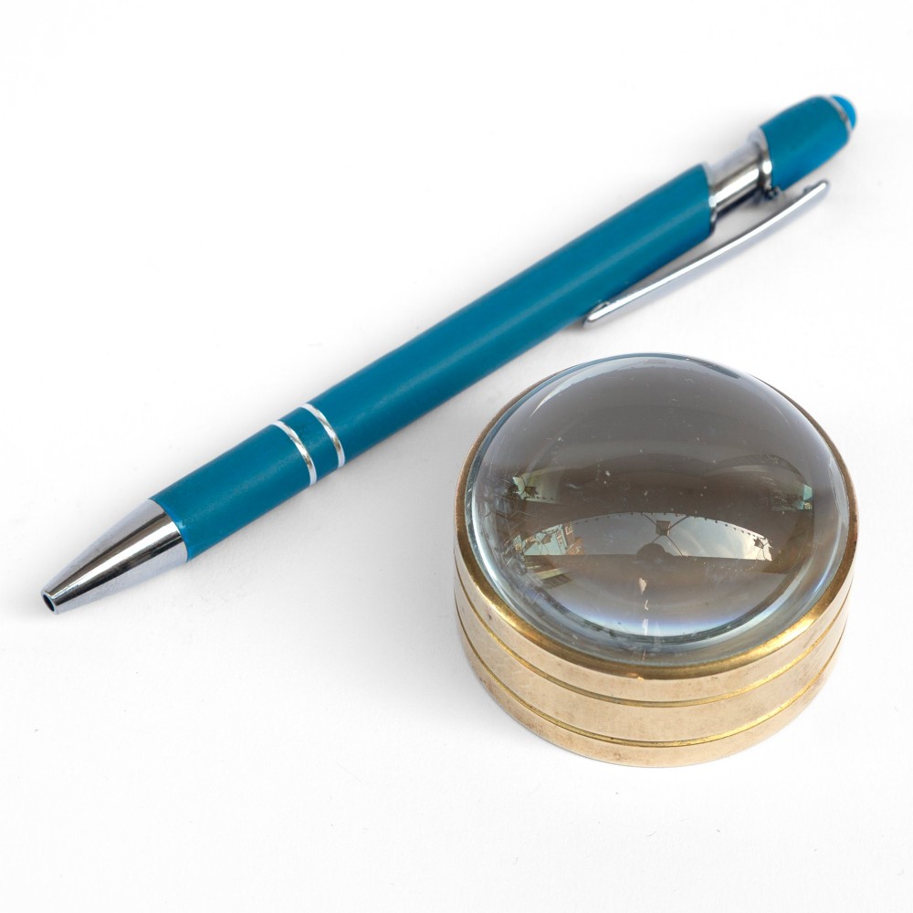 A solid circular brass mounted glass dome desk magnifying lens shown on a white background.