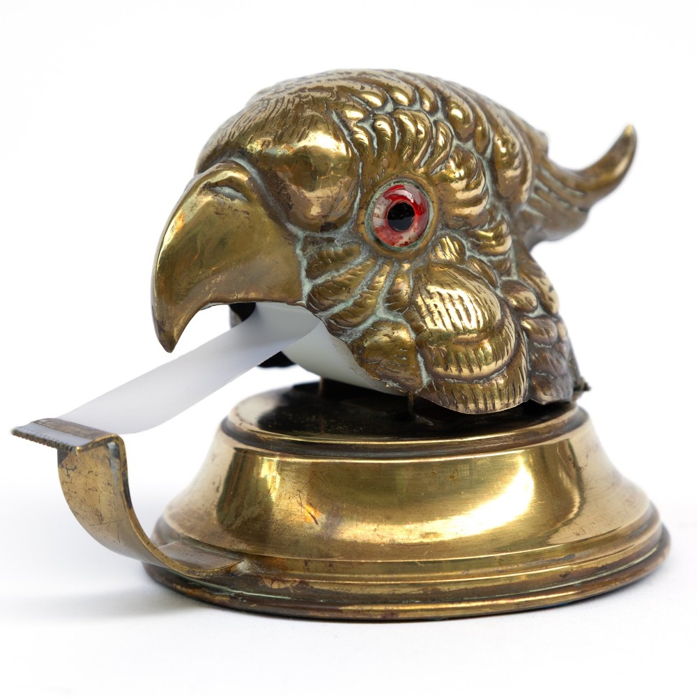 A cast brass desk tape dispenser formed in the shape of a parrot head with red glass eyes set on a round stepped base.