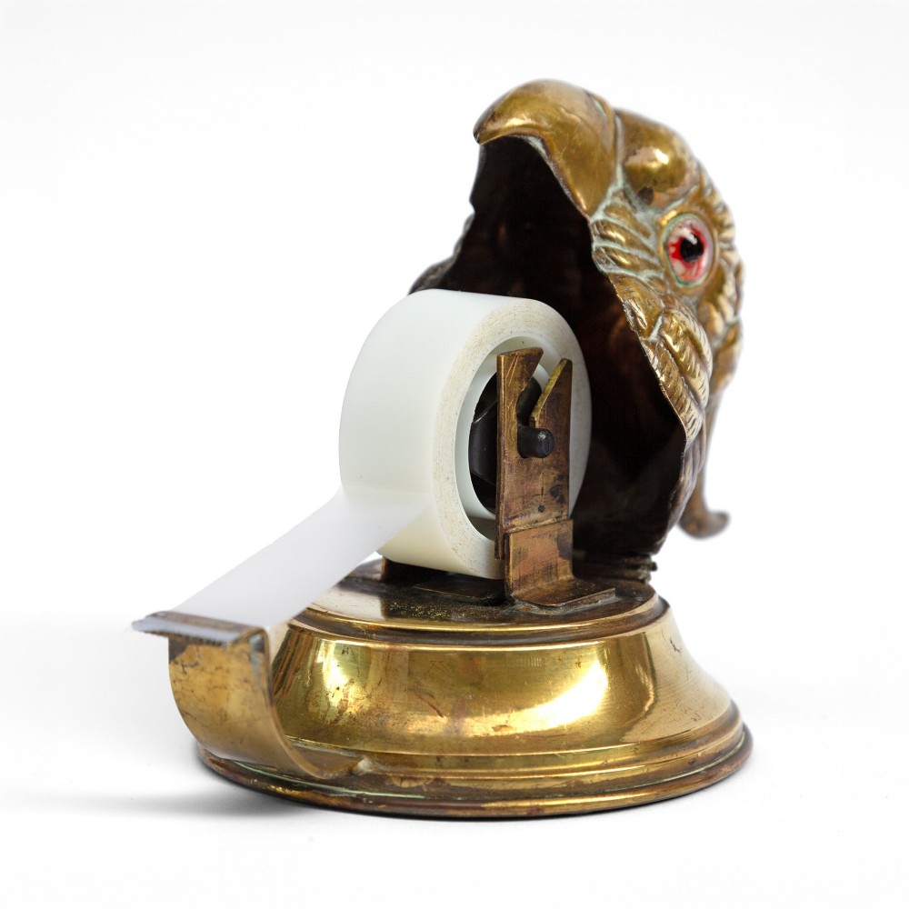 A cast brass desk tape dispenser formed in the shape of a parrot head with red glass eyes set on a round stepped base. View VI