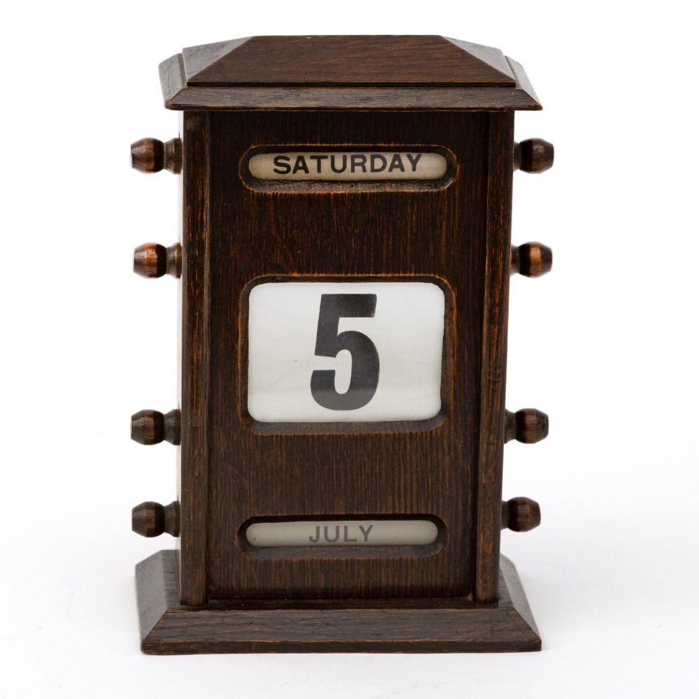 A dark oak desktop perpetual calendar with turned side knobs and apertures displaying the day, date, and month. View II