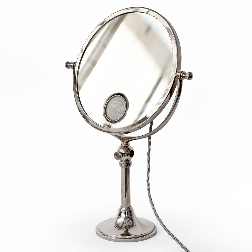 A nickel-plated circular vanity mirror featuring an adjustable swivel stem, circular weighted base, integrated central lens, and braided electrical cord.