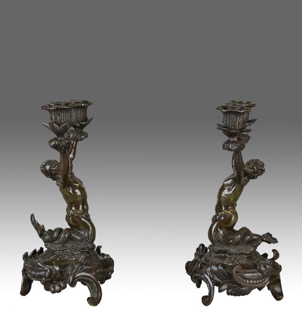 A pair of dark patinated bronze candlesticks shaped as mythical mermen raising leafy candle sconces on decorative scrolled bases.
