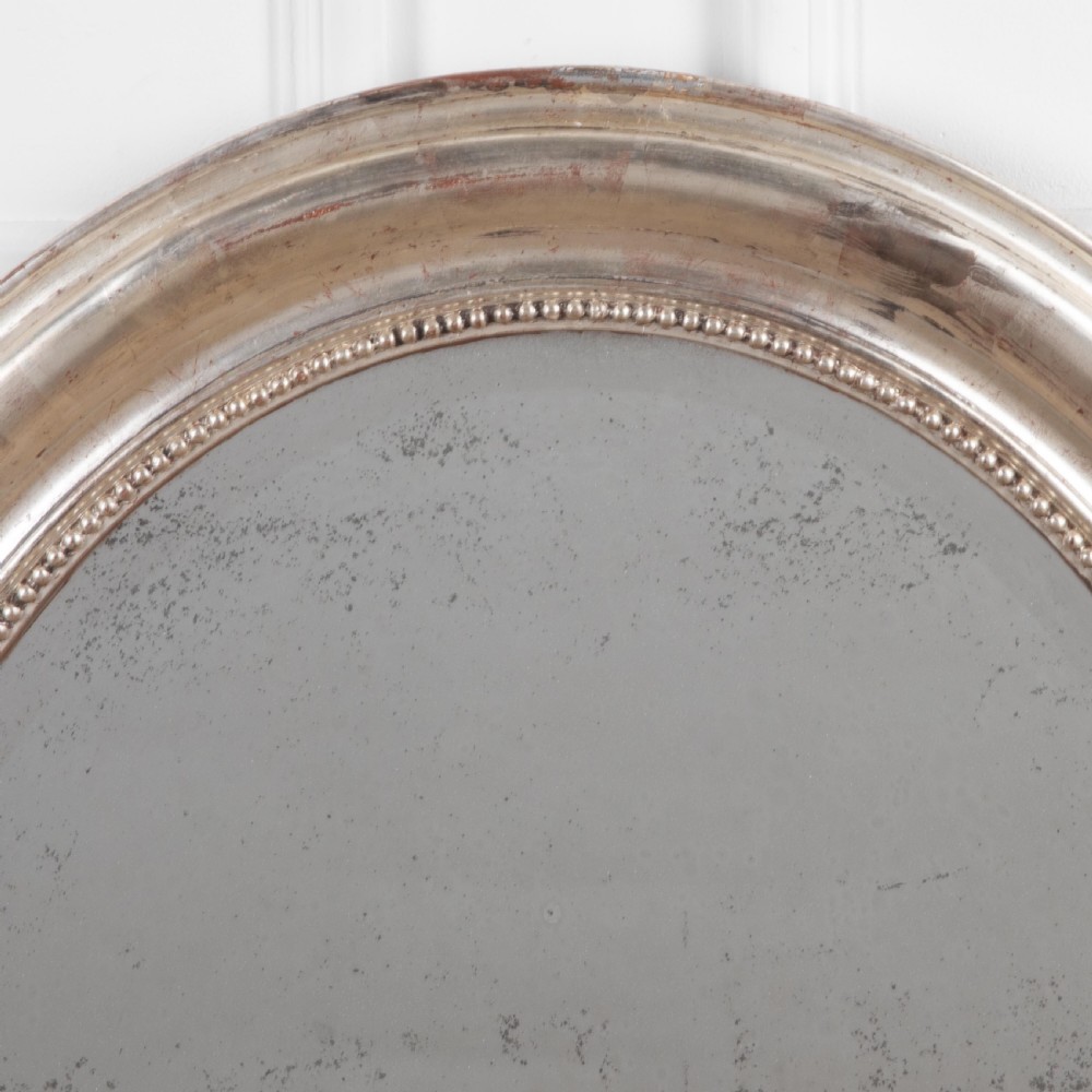 A 19th-century oval wall mirror featuring a silvered moulding frame with beaded rim detail and aged glass showing natural foxing. View VI
