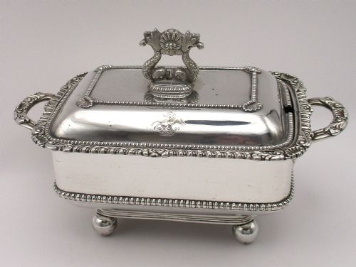 fig 1. A rectangular antique silver-plated sauce tureen with an engraved crest, decorative dolphin finial, gadrooned trim, twin handles, and ball feet.