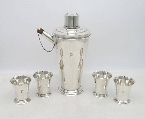 fig 1. An Art Deco silver-plated cocktail set consisting of a tapered shaker with a spout and chain-attached stopper alongside four matching small cups, all engraved with the initial P.