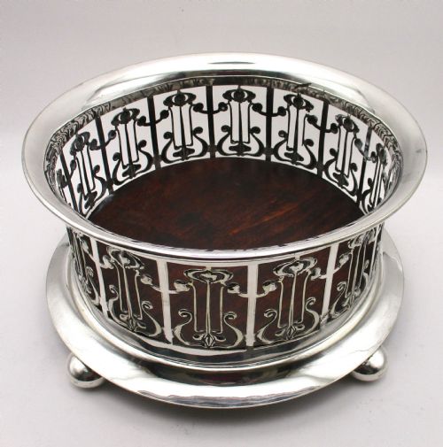 fig 1. A circular Art Nouveau silver-plated coaster featuring openwork pierced floral fretwork around the gallery and a polished dark wood base resting on four small ball feet.