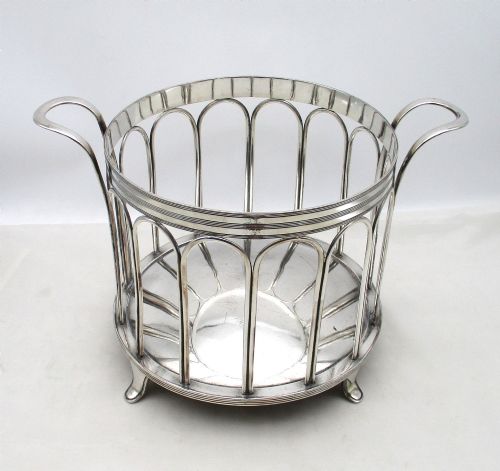 fig 1. A silver-plated cylindrical bottle coaster featuring arched wirework sides, twin looped handles, a reeded rim, and small curved feet.
