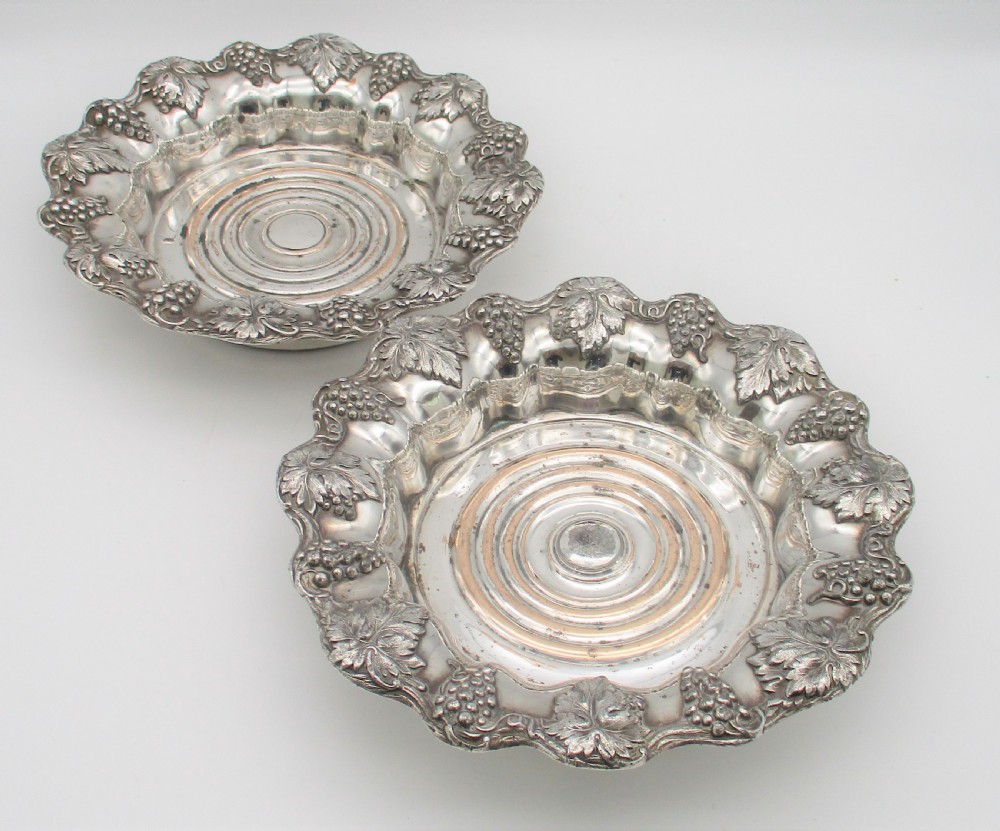 A pair of circular silver-plated wine coasters featuring scalloped rims decorated with grapevine and foliage relief details around concentric circular turned bases. View II
