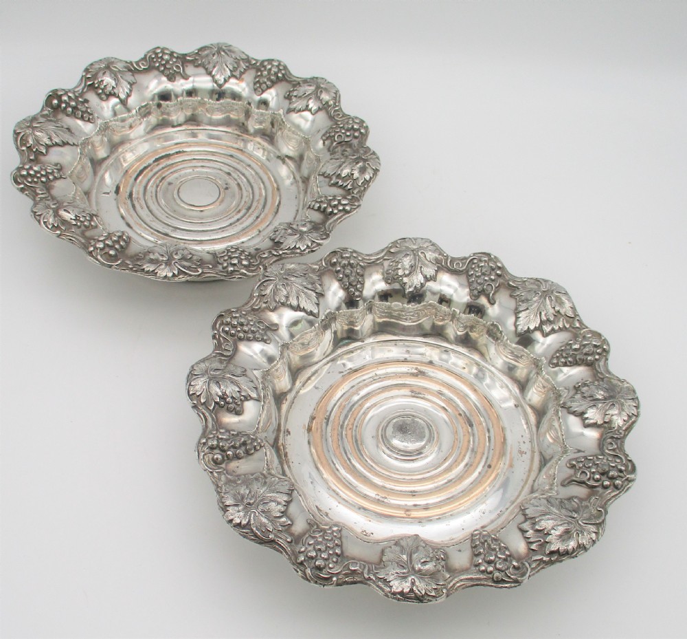 A pair of circular silver-plated wine coasters featuring scalloped rims decorated with grapevine and foliage relief details around concentric circular turned bases. View IV