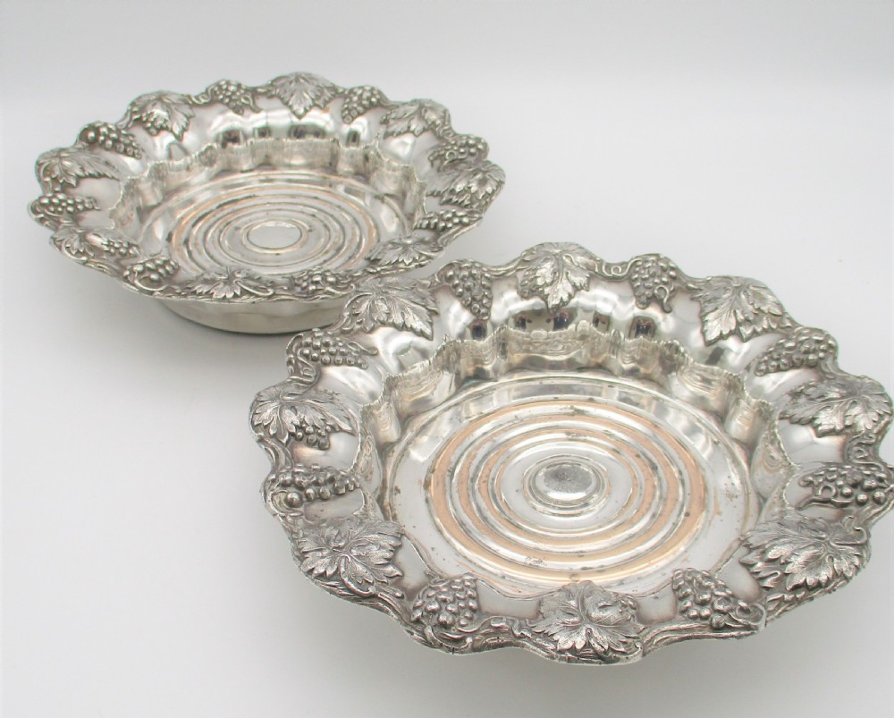 A pair of circular silver-plated wine coasters featuring scalloped rims decorated with grapevine and foliage relief details around concentric circular turned bases. View V