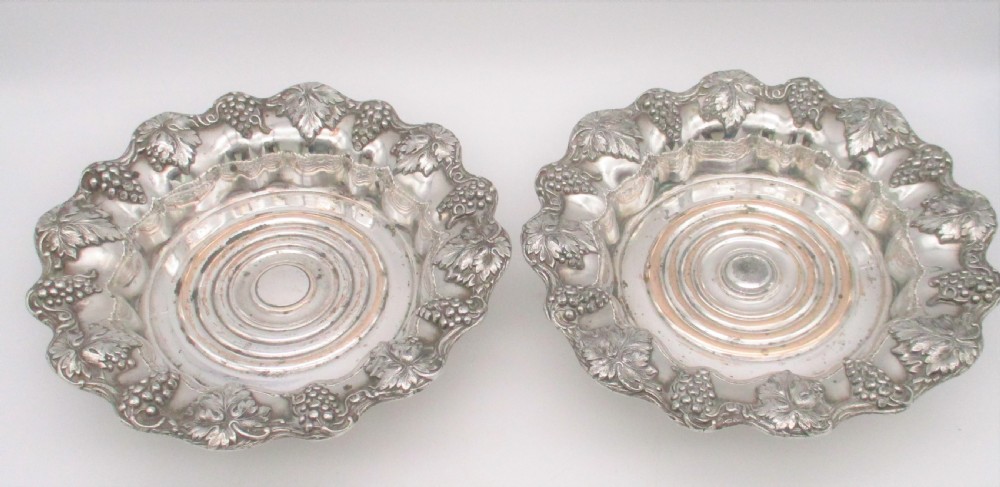 A pair of circular silver-plated wine coasters featuring scalloped rims decorated with grapevine and foliage relief details around concentric circular turned bases. View VI