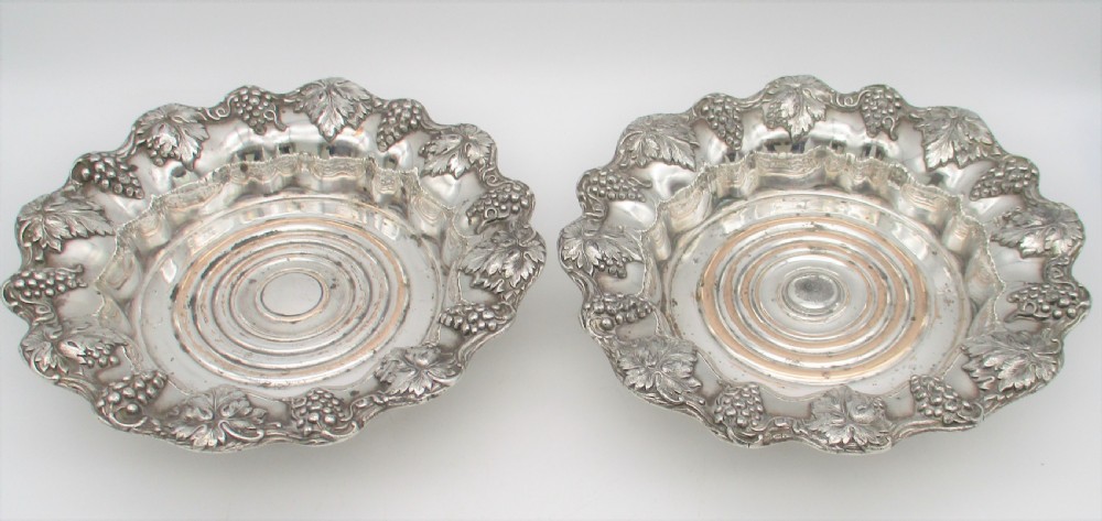 A pair of circular silver-plated wine coasters featuring scalloped rims decorated with grapevine and foliage relief details around concentric circular turned bases. View VII