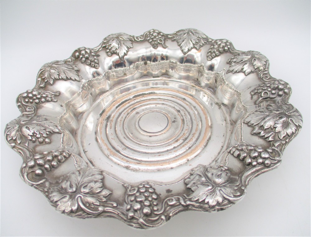A pair of circular silver-plated wine coasters featuring scalloped rims decorated with grapevine and foliage relief details around concentric circular turned bases. View X