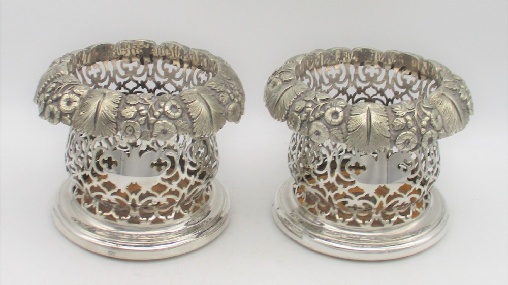 A pair of mid-19th-century silver-plated wine bottle coasters featuring pierced lattice sides, floral repousse borders, and turned wooden bases.