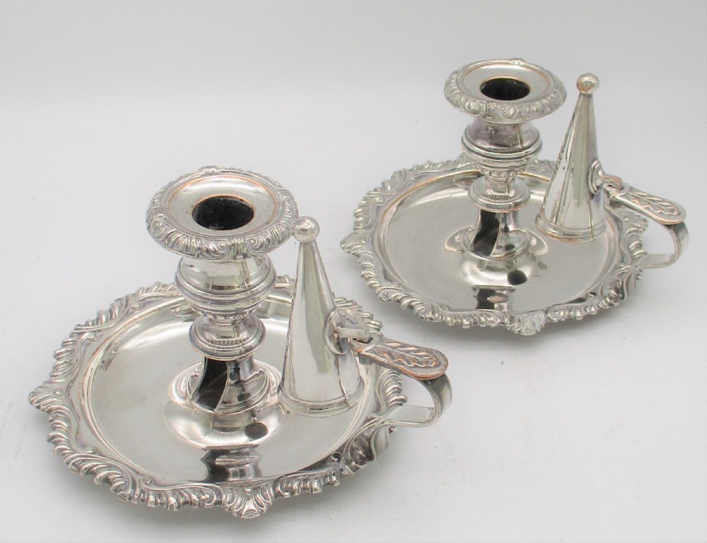 A pair of silver-plated chamber candlesticks featuring wide circular scroll-rimmed bases, integrated finger handles, and removable conical candle snuffers.