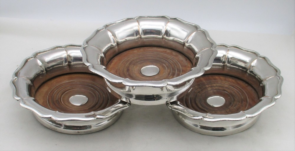 A set of three silver-plated wine bottle coasters with scalloped rims and turned wood bases.