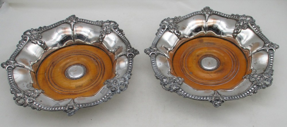 A pair of circular Regency silver-plated decanter coasters featuring turned wooden bases, central silver bosses, and ornate shell and scroll borders.