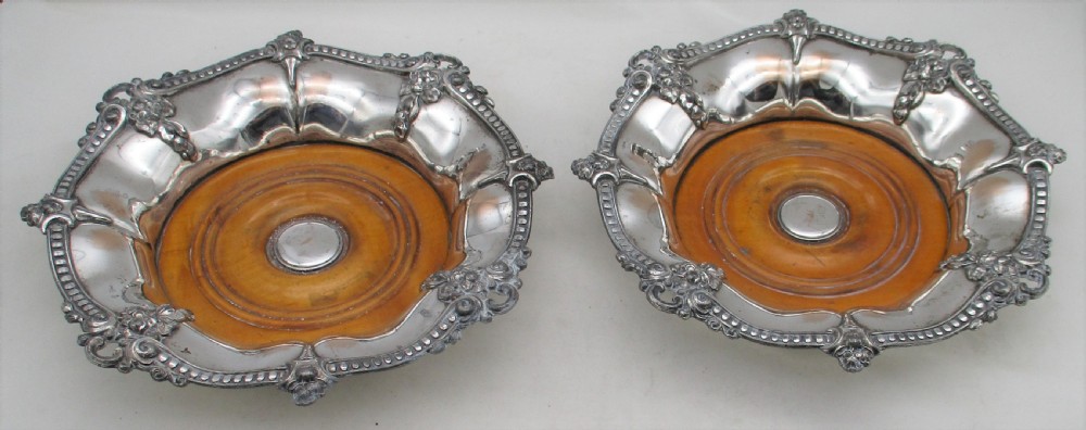 A pair of circular Regency silver-plated decanter coasters featuring turned wooden bases, central silver bosses, and ornate shell and scroll borders. View II