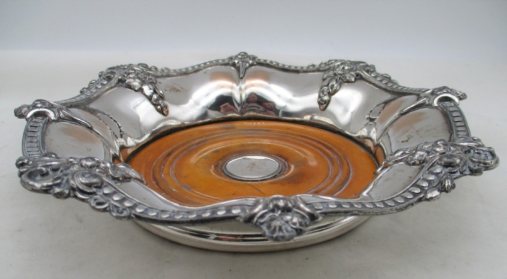 A pair of circular Regency silver-plated decanter coasters featuring turned wooden bases, central silver bosses, and ornate shell and scroll borders. View V