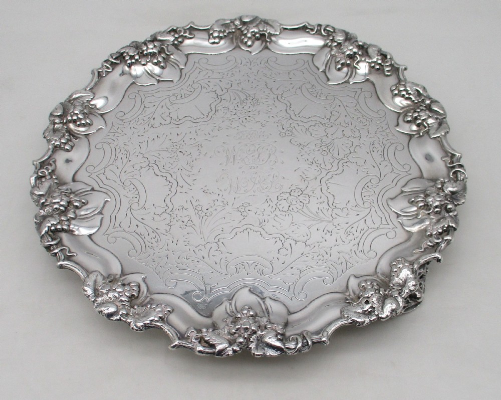 A circular silver-plated tray featuring engraved scrollwork across the surface and a raised border decorated with molded grapevine and foliage motifs.