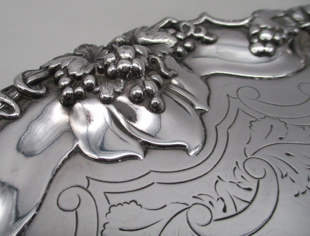 A circular silver-plated tray featuring engraved scrollwork across the surface and a raised border decorated with molded grapevine and foliage motifs. View V