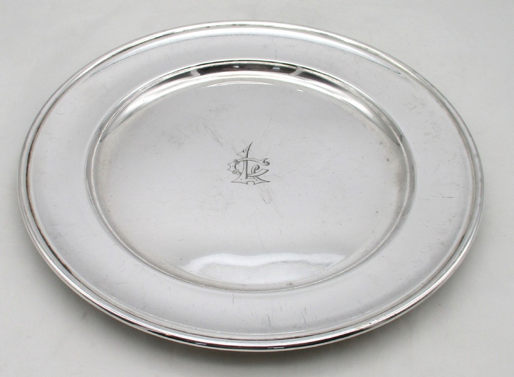 A set of four circular silver plated charger plates featuring wide flat rims and engraved central inscriptions and monograms. View IV