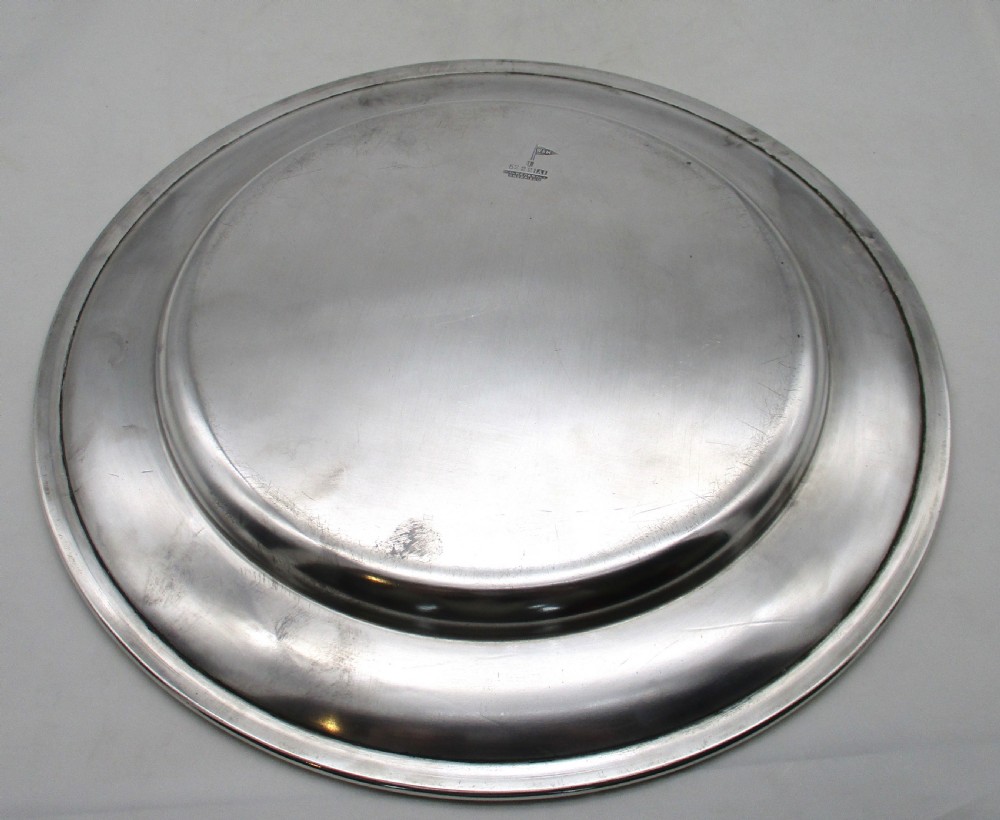 A set of four circular silver plated charger plates featuring wide flat rims and engraved central inscriptions and monograms. View VI