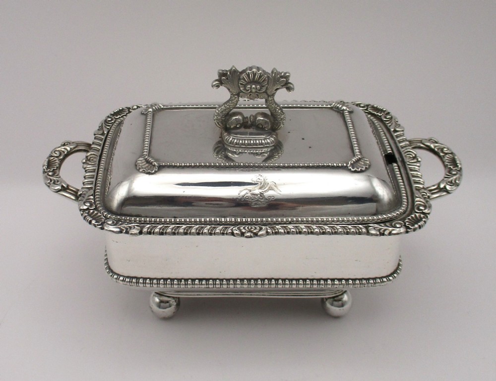 A rectangular antique silver-plated sauce tureen with an engraved crest, decorative dolphin finial, gadrooned trim, twin handles, and ball feet.