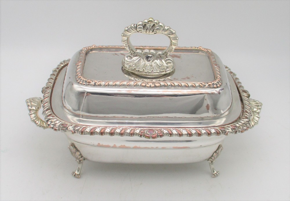 An antique rectangular silver-plated sauce tureen with a fitted lid, gadrooned borders, a shell-shaped handle, and cast decorative feet showing copper exposure.