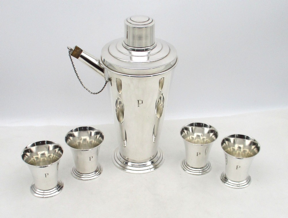 An Art Deco silver-plated cocktail set consisting of a tapered shaker with a spout and chain-attached stopper alongside four matching small cups, all engraved with the initial P.