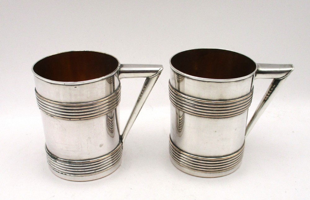 A pair of cylindrical silver-plated tankards featuring horizontal reeded bands, angular handles, and gilded interiors.