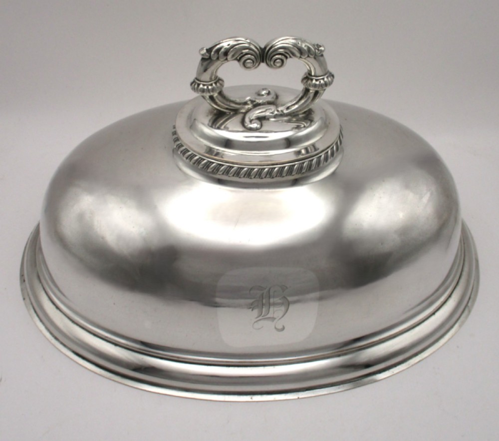 An oval domed silver-plated food cover featuring a cast leaf motif top handle and an engraved crest on the front.