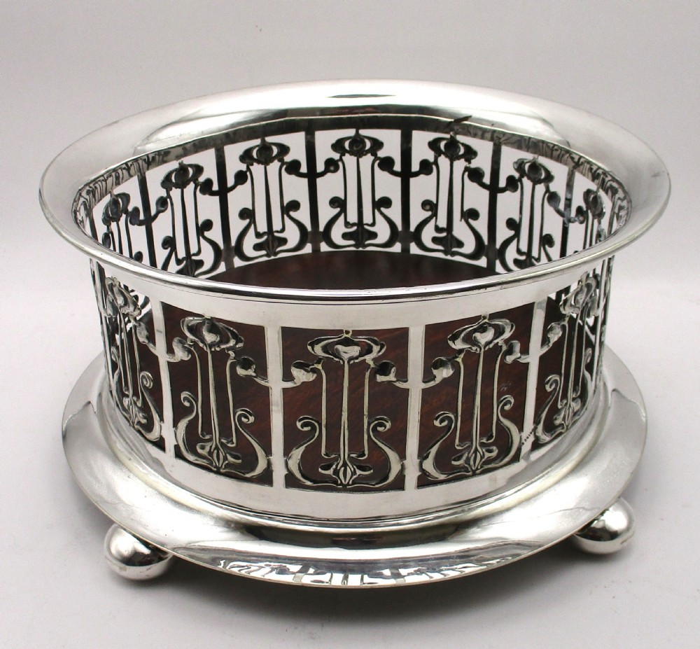 A circular Art Nouveau silver-plated coaster featuring openwork pierced floral fretwork around the gallery and a polished dark wood base resting on four small ball feet.
