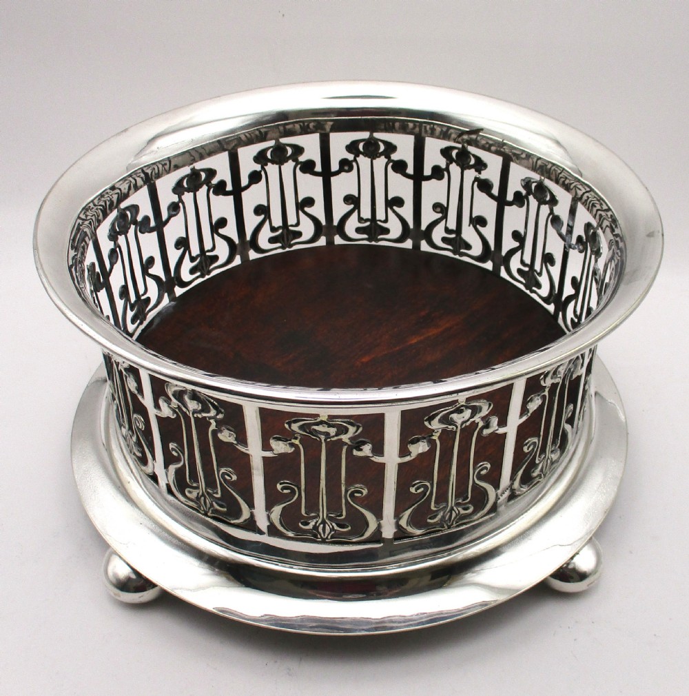A circular Art Nouveau silver-plated coaster featuring openwork pierced floral fretwork around the gallery and a polished dark wood base resting on four small ball feet. View II