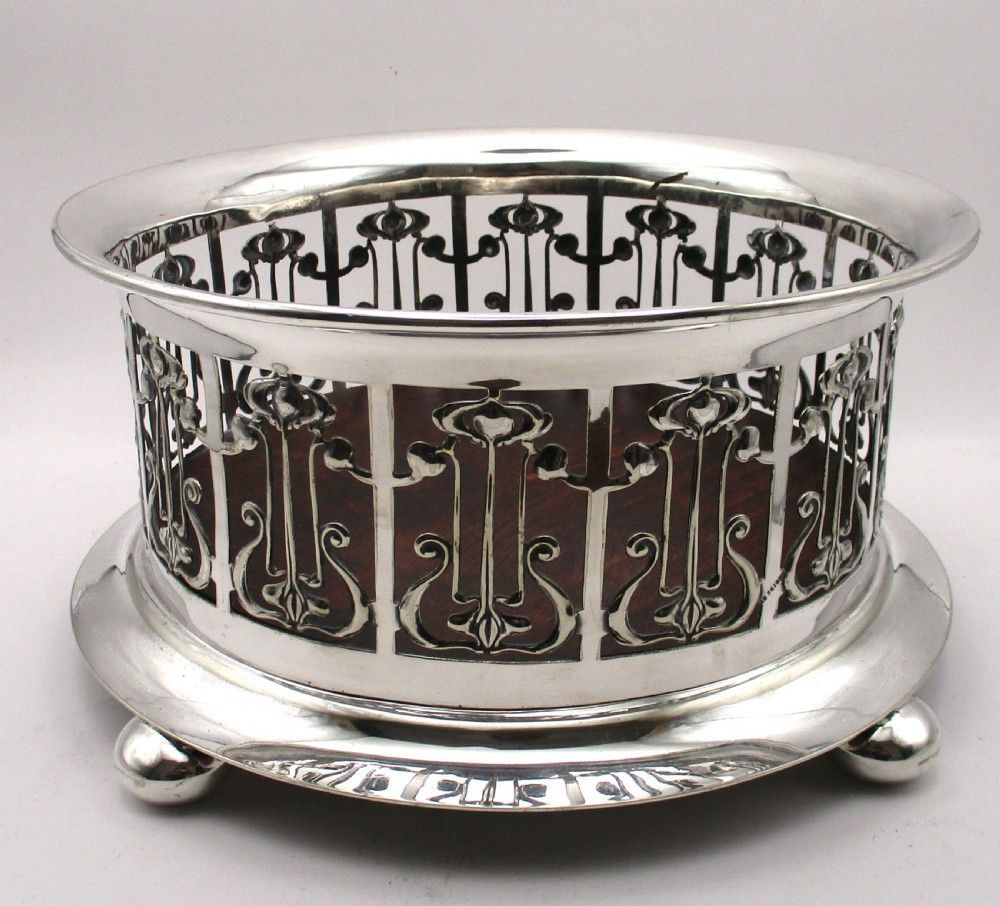 A circular Art Nouveau silver-plated coaster featuring openwork pierced floral fretwork around the gallery and a polished dark wood base resting on four small ball feet. View III