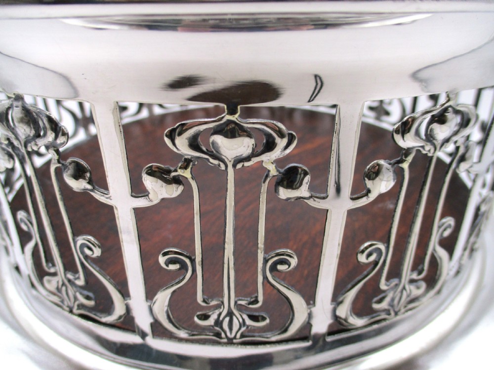 A circular Art Nouveau silver-plated coaster featuring openwork pierced floral fretwork around the gallery and a polished dark wood base resting on four small ball feet. View IV