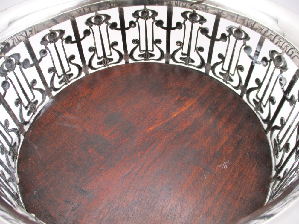 A circular Art Nouveau silver-plated coaster featuring openwork pierced floral fretwork around the gallery and a polished dark wood base resting on four small ball feet. View V