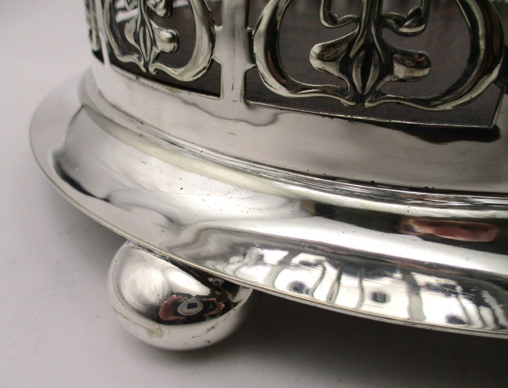 A circular Art Nouveau silver-plated coaster featuring openwork pierced floral fretwork around the gallery and a polished dark wood base resting on four small ball feet. View VI