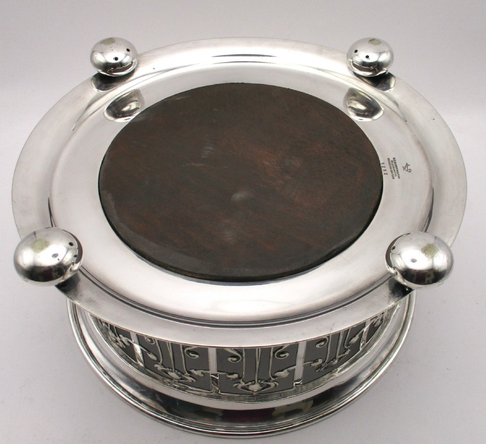 A circular Art Nouveau silver-plated coaster featuring openwork pierced floral fretwork around the gallery and a polished dark wood base resting on four small ball feet. View VII