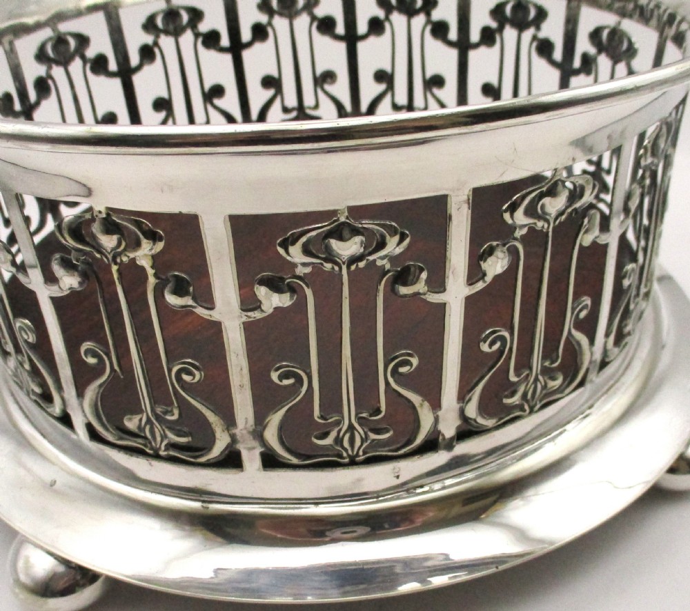 A circular Art Nouveau silver-plated coaster featuring openwork pierced floral fretwork around the gallery and a polished dark wood base resting on four small ball feet. View IX