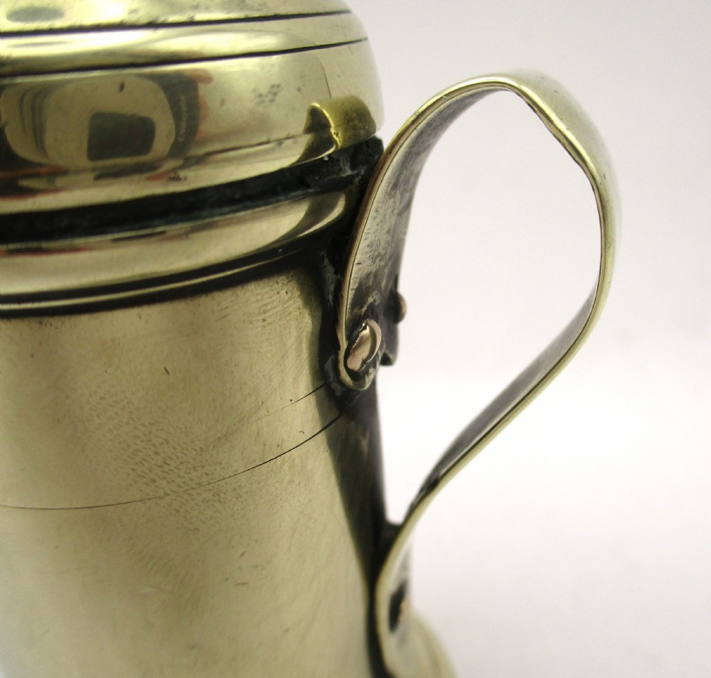 An eighteenth-century brass spice dredger featuring a perforated domed lid, a cylindrical body with moulded bands, a curved strap handle, and a stepped circular base. View V