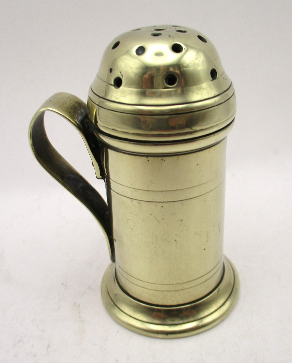 An eighteenth-century brass spice dredger featuring a perforated domed lid, a cylindrical body with moulded bands, a curved strap handle, and a stepped circular base. View VII