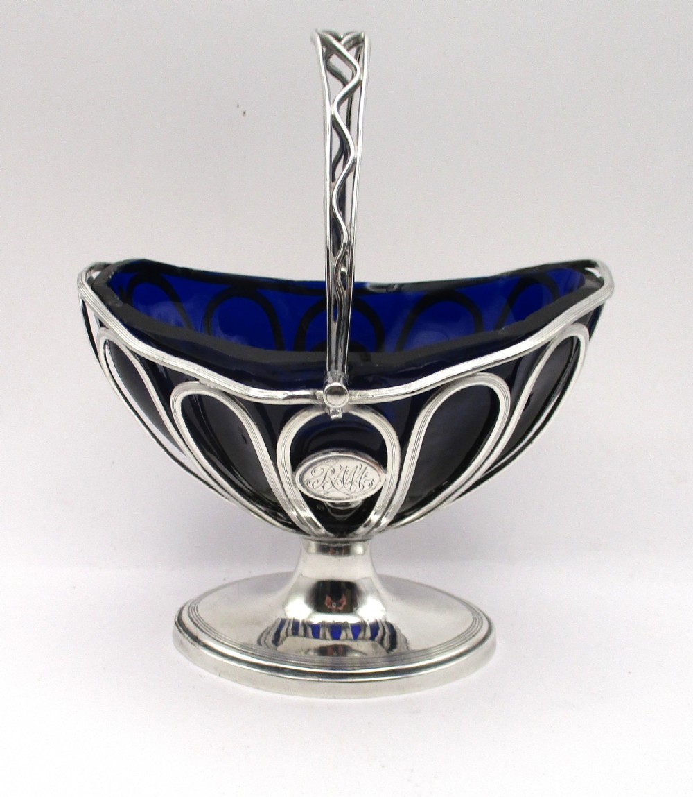 A Georgian Sheffield plated silver wirework sugar basket featuring an oval engraved cartouche, overhead handle, pedestal foot, and cobalt blue glass liner. View II