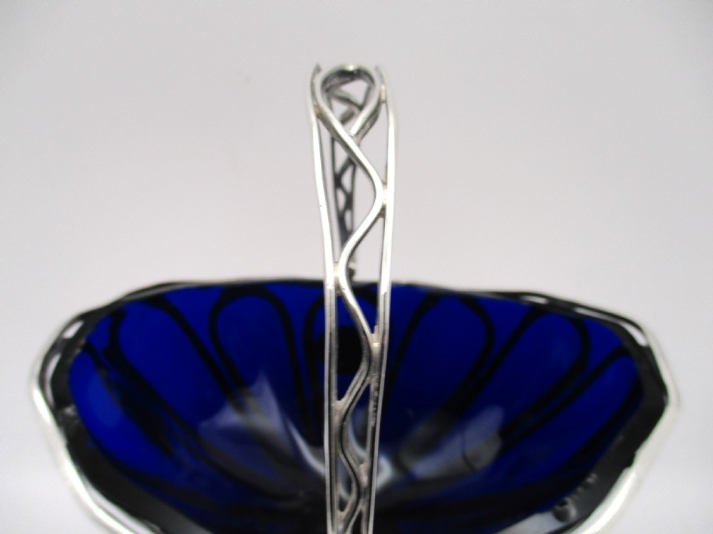 A Georgian Sheffield plated silver wirework sugar basket featuring an oval engraved cartouche, overhead handle, pedestal foot, and cobalt blue glass liner. View IV