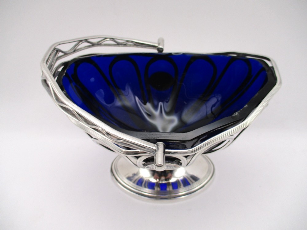 A Georgian Sheffield plated silver wirework sugar basket featuring an oval engraved cartouche, overhead handle, pedestal foot, and cobalt blue glass liner. View VI