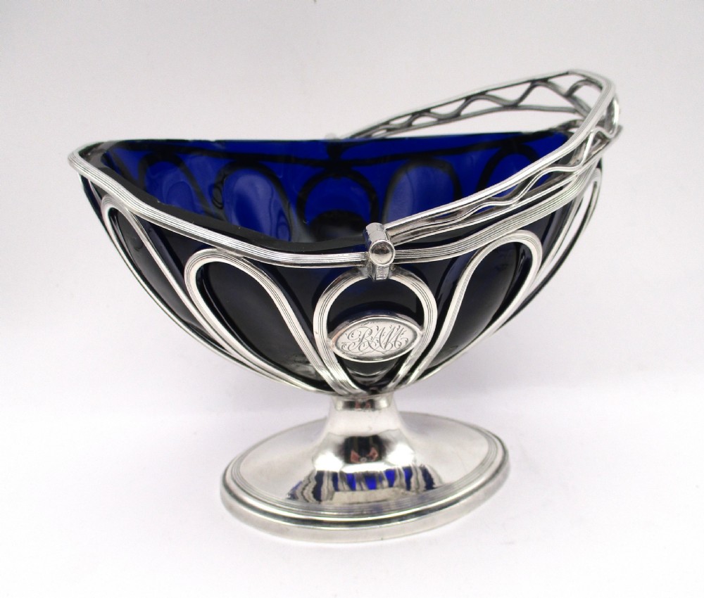 A Georgian Sheffield plated silver wirework sugar basket featuring an oval engraved cartouche, overhead handle, pedestal foot, and cobalt blue glass liner. View VII