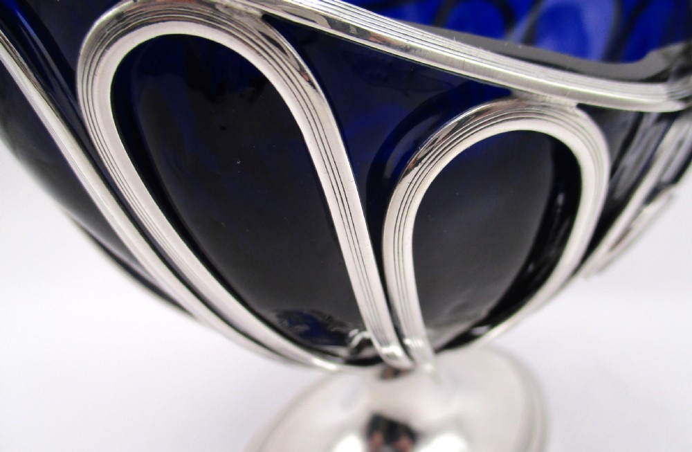A Georgian Sheffield plated silver wirework sugar basket featuring an oval engraved cartouche, overhead handle, pedestal foot, and cobalt blue glass liner. View VIII